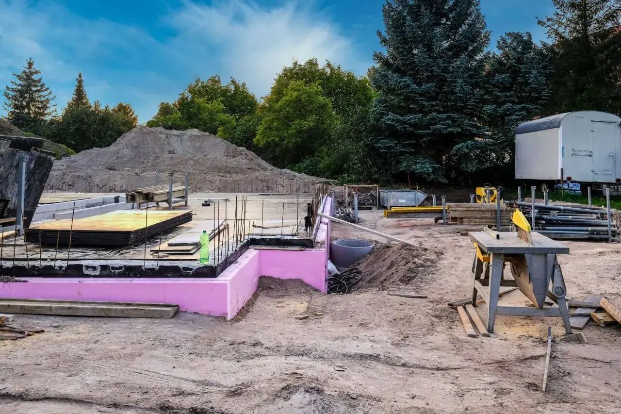 Budowa w stanie zero: fundamenty z różową izolacją, zbrojenie, sterta piasku i sprzęt budowlany.