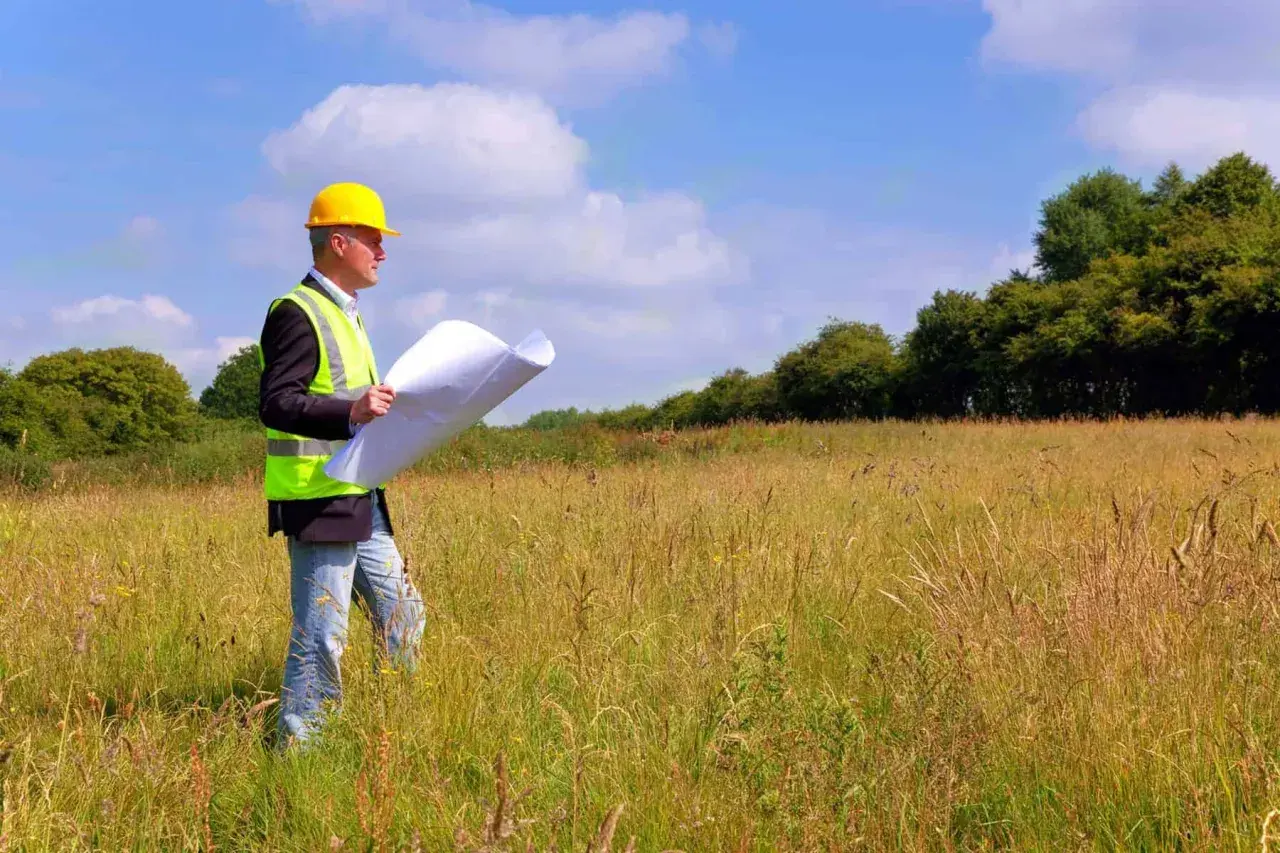 Inżynier w kasku i kamizelce odblaskowej analizuje plany podczas badań geotechnicznych gruntu na łące.