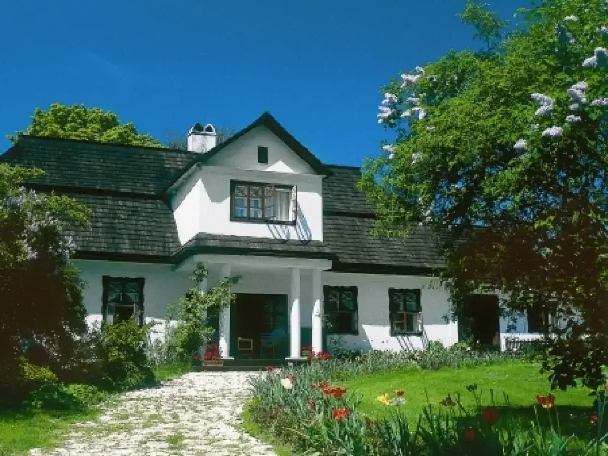 Piękny biały dom z ciemnym dachem, otoczony zielenią i kwitnącymi kwiatami. Widać ścieżkę z kamieni i ganek, jakby zrealizowano dobudowa ganku projekt.