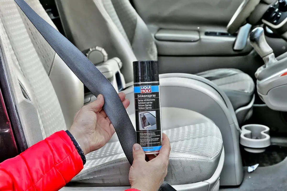 Ręce trzymające pas bezpieczeństwa i puszkę z napisem "Silikonspray". Preparat ten to olej silikonowy do paneli, który ułatwia konserwację wnętrza samochodu.