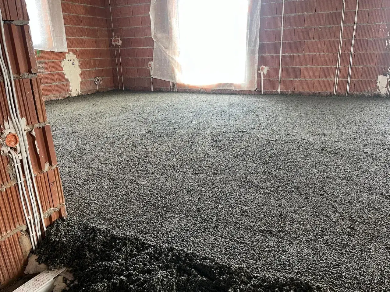 Świeżo wylany styrobeton na podłodze w budowanym pomieszczeniu. Widać ceglane ściany i okna.