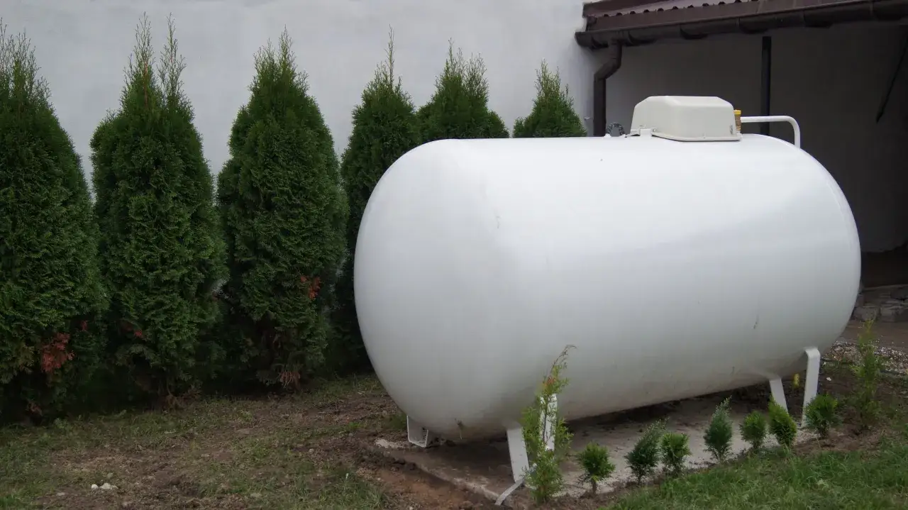 Biały, poziomy zbiornik na gaz płynny stoi na trawniku, otoczony żywopłotem z cyprysów.