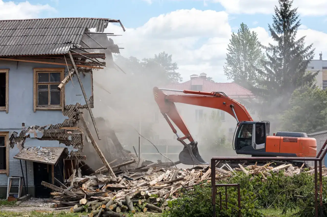 Pomarańczowy koparka prowadzi rozbiórkę budynku gospodarczego, wzbijając tumany kurzu. Wszędzie widać gruzy i drewno.