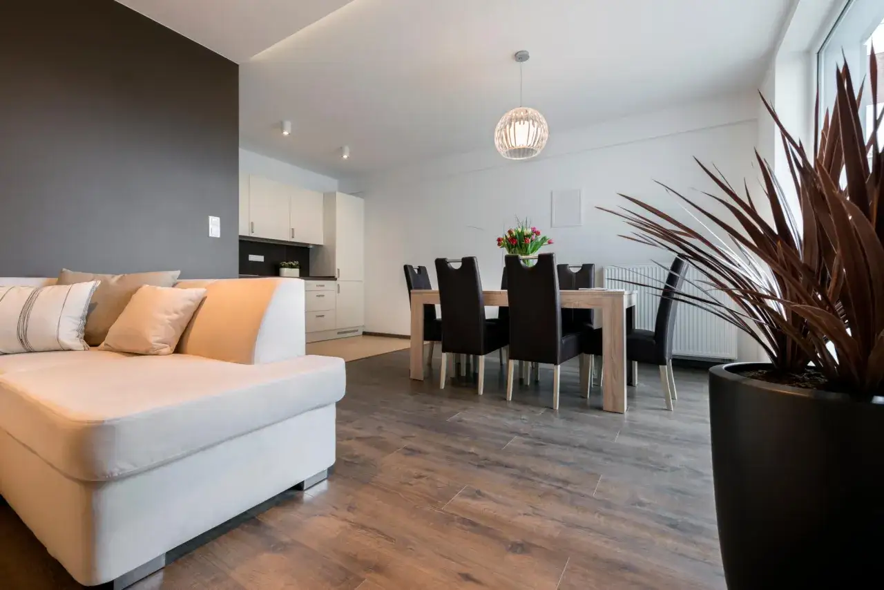 Jasny salon z jadalnią i kuchnią, gdzie standardowa wysokość mieszkania pozwala na swobodę aranżacji. Wygodna sofa, stół z krzesłami i duża roślina.