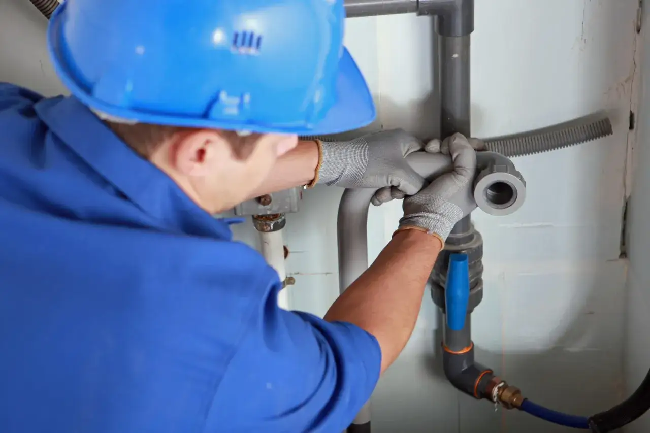 Instalator w niebieskim kombinezonie i kasku montuje rury, prawdopodobnie rozwiązując problem braku odpowietrzenia pionu.
