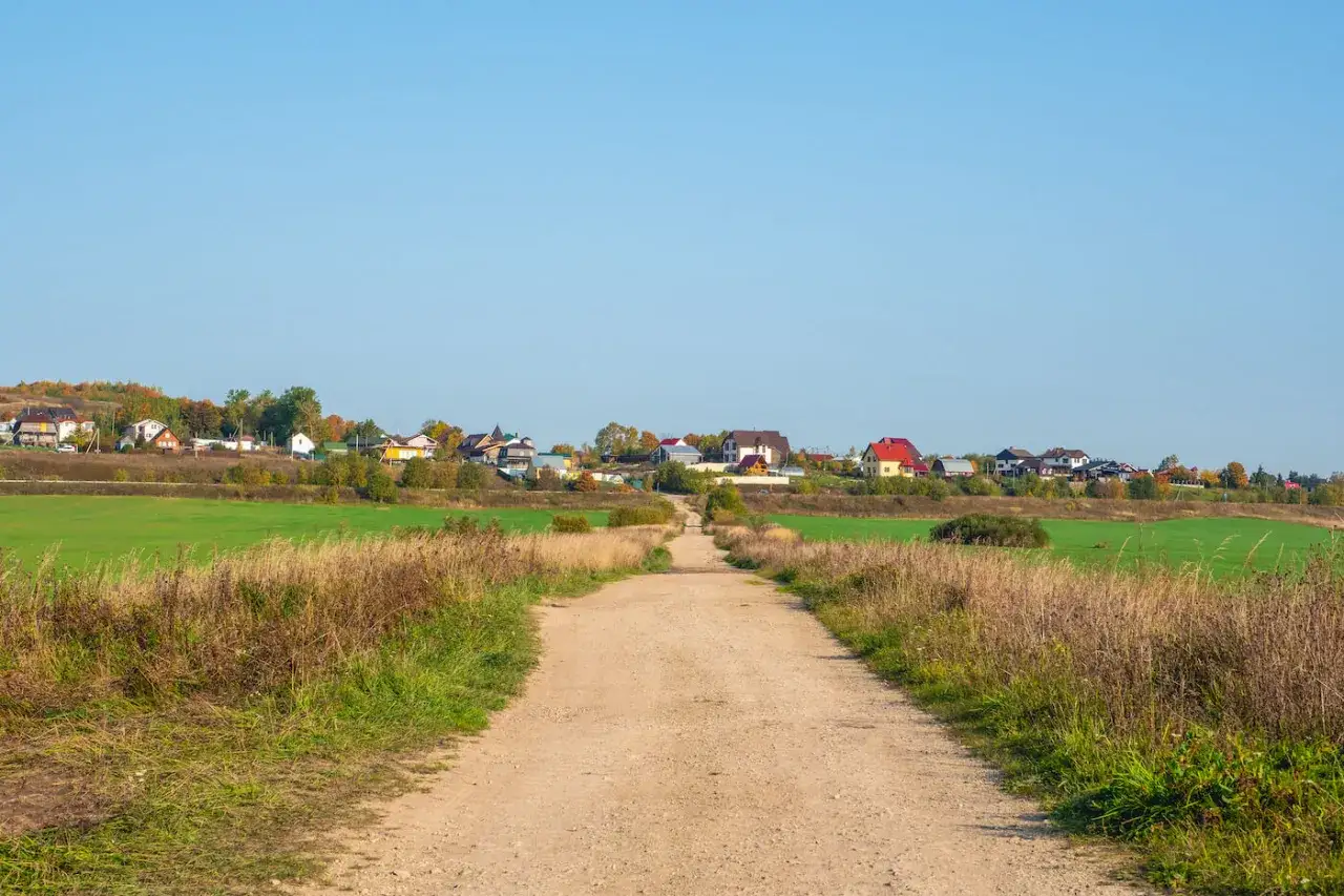 Droga gruntowa prowadzi do osady domków jednorodzinnych. Minimalna szerokość drogi dojazdowej do posesji jest widoczna na tle zielonych pól i błękitnego nieba.