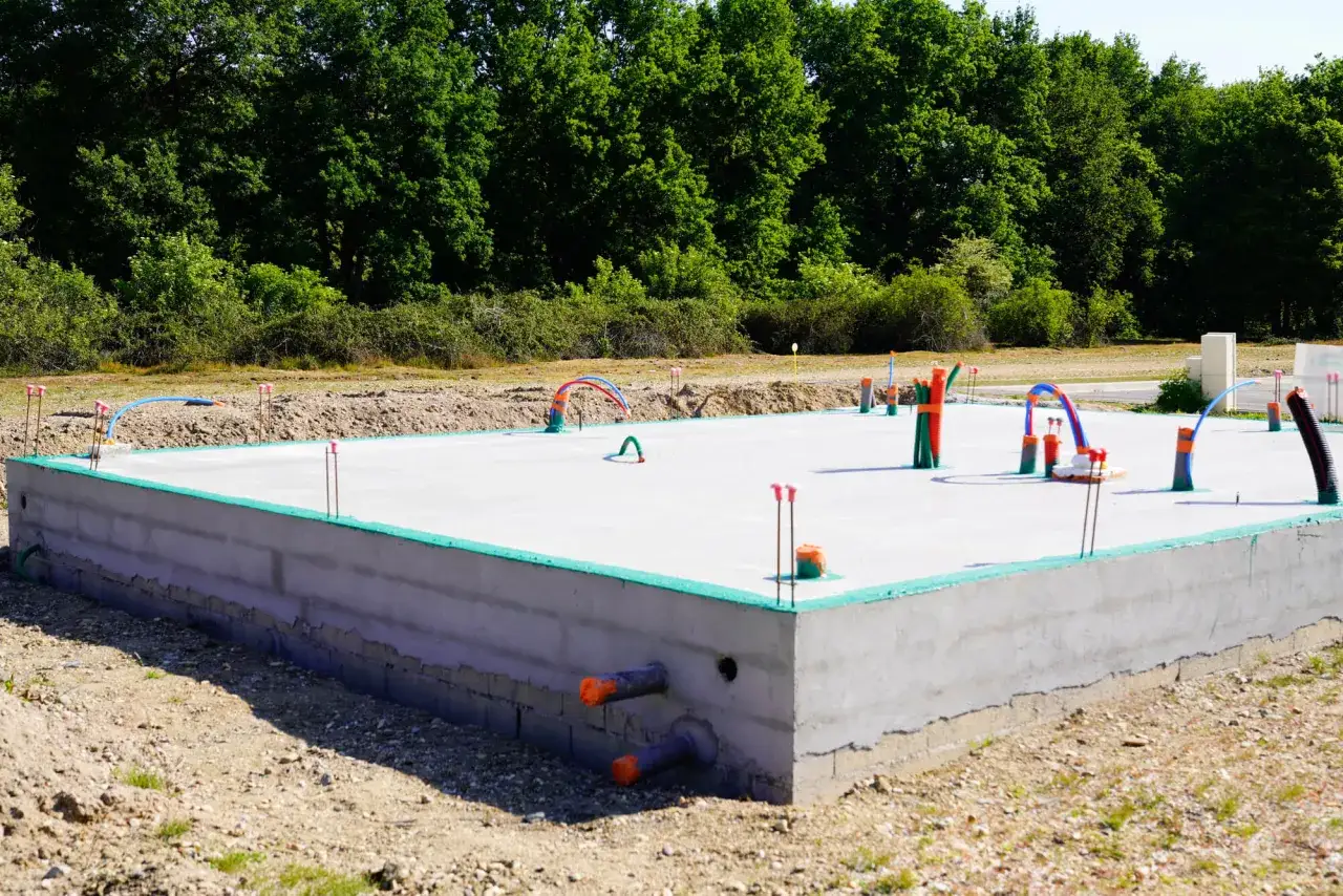 Gotowa płyta fundamentowa pod garaż z instalacją hydrauliczną i elektryczną, otoczona zieloną izolacją i betonowym murem.