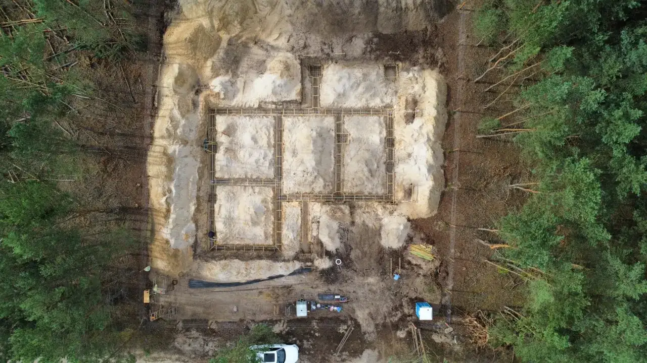 Budowa fundamentów w lesie. Widać zbrojenie i szalunki, przygotowane do betonowania. W tle drzewa i sprzęt budowlany. To etap prac poprzedzający właściwe badania geotechniczne gruntu.