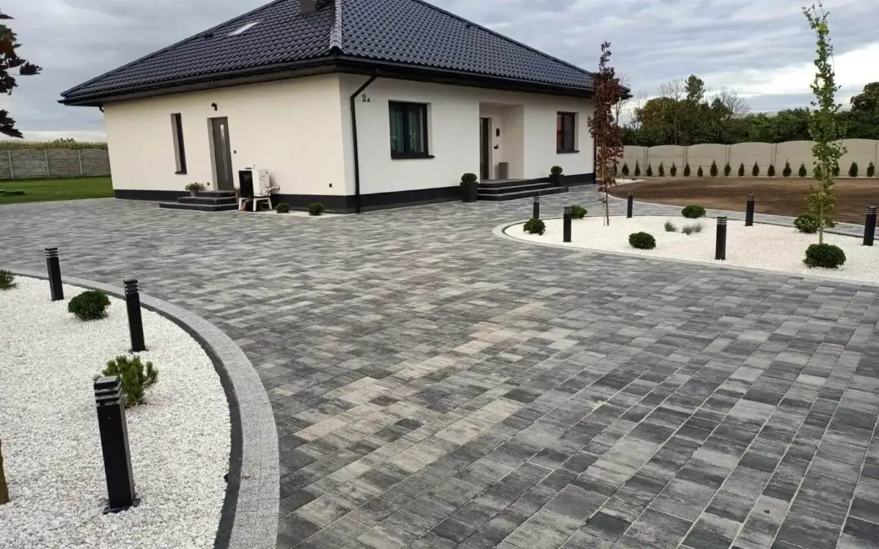 Nowoczesny podjazd z kostki brukowej prowadzi do białego domu. Oświetlenie i biały grys podkreślają jego elegancję.