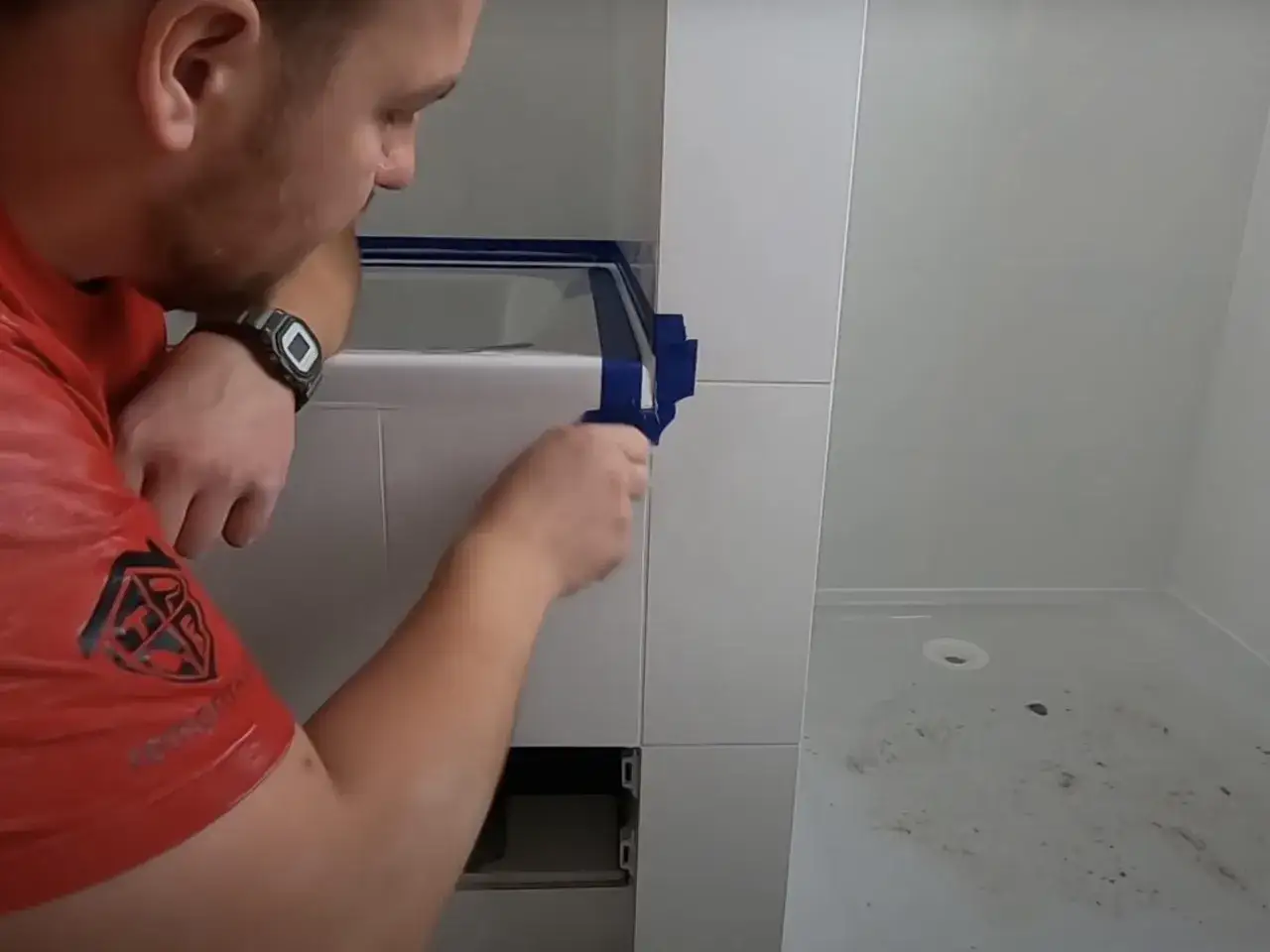 Mężczyzna w czerwonej koszulce przykleja taśmę, przygotowując się do tego, jak położyć silikon w narożnikach łazienki.