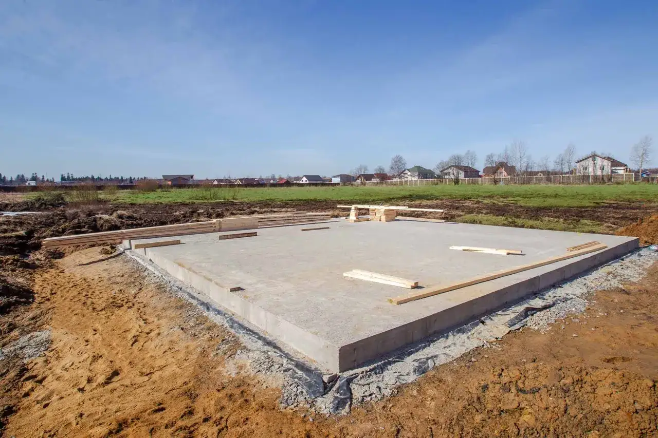Betonowa płyta fundamentowa, gotowa pod budowę domu. W tle widać inne domy i zielone pola.
