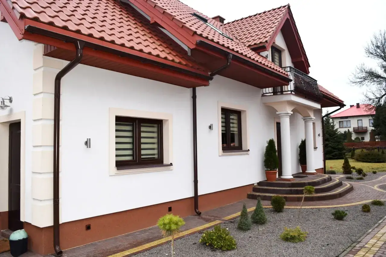 Biały dom z brązowym cokołem i czerwonym dachem. Wokół zadbany ogród z kamieniami i iglakami.