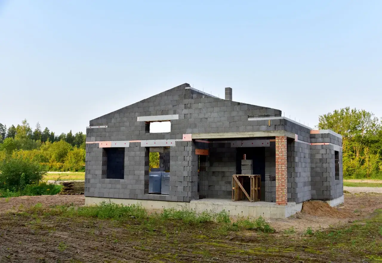 Budowa domu z bloczków, które mogą być wykonane z keramzytu. Widać fundamenty, ściany z otworami na okna i drzwi, oraz komin.
