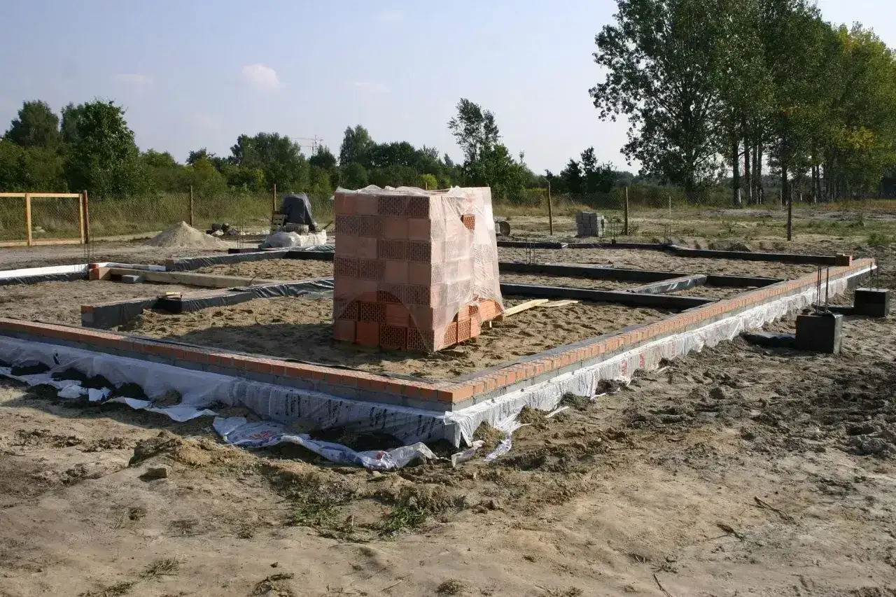 Stan zero budynku: fundamenty z cegły, piasek, folia i stos materiałów budowlanych.