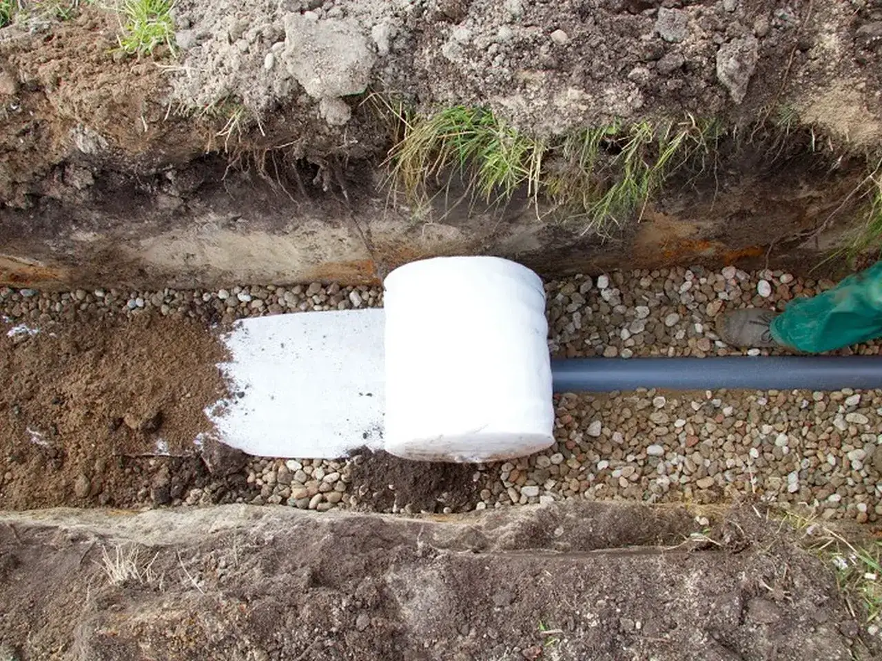 Instalacja drenażu: rura drenarska owinięta geowłókniną, otoczona żwirem i ziemią, gotowa do zasypania.