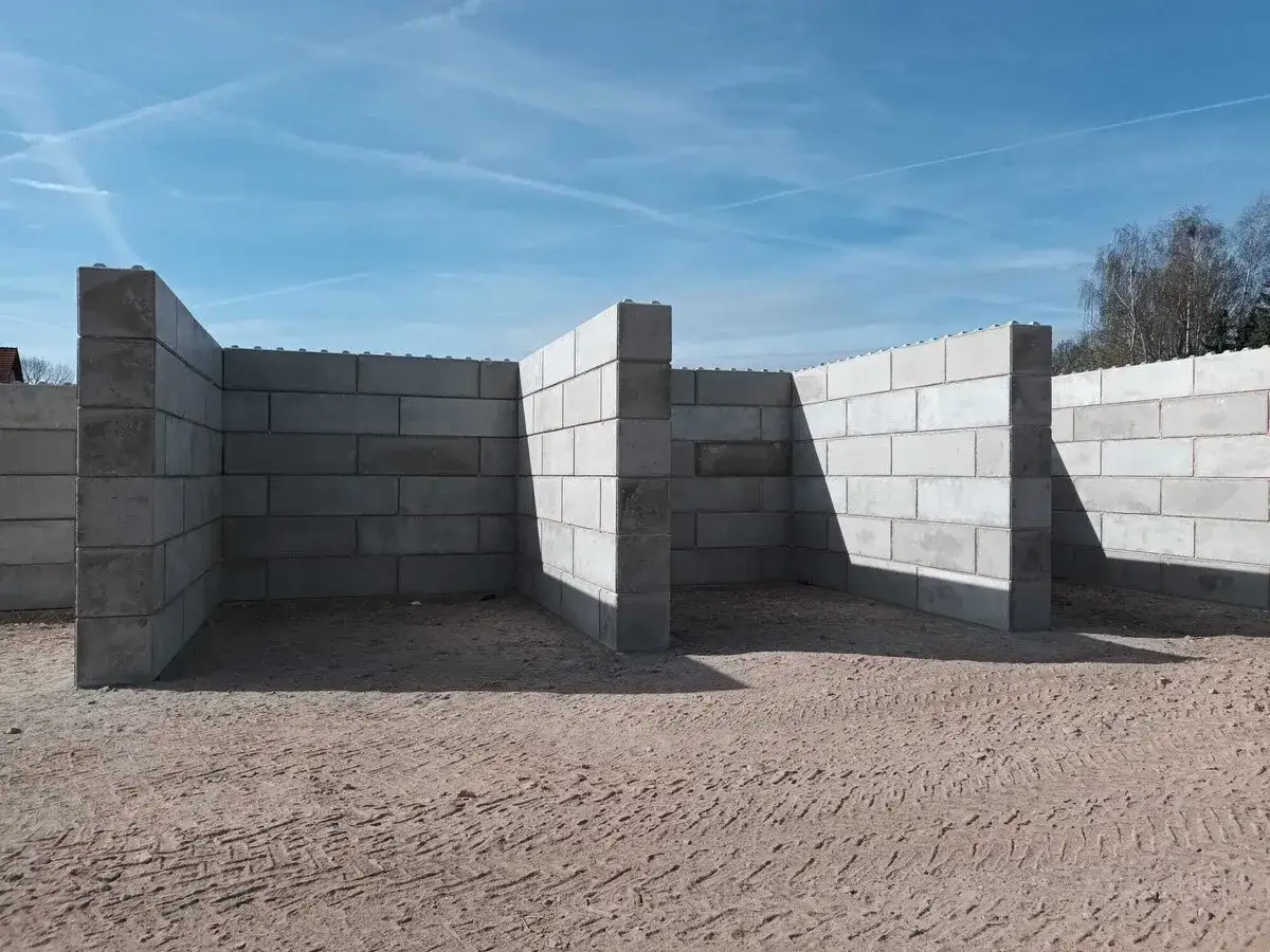 Budowa muru oporowego z bloczków betonowych. Konstrukcja tworzy zarys pomieszczenia na piaszczystym placu budowy.