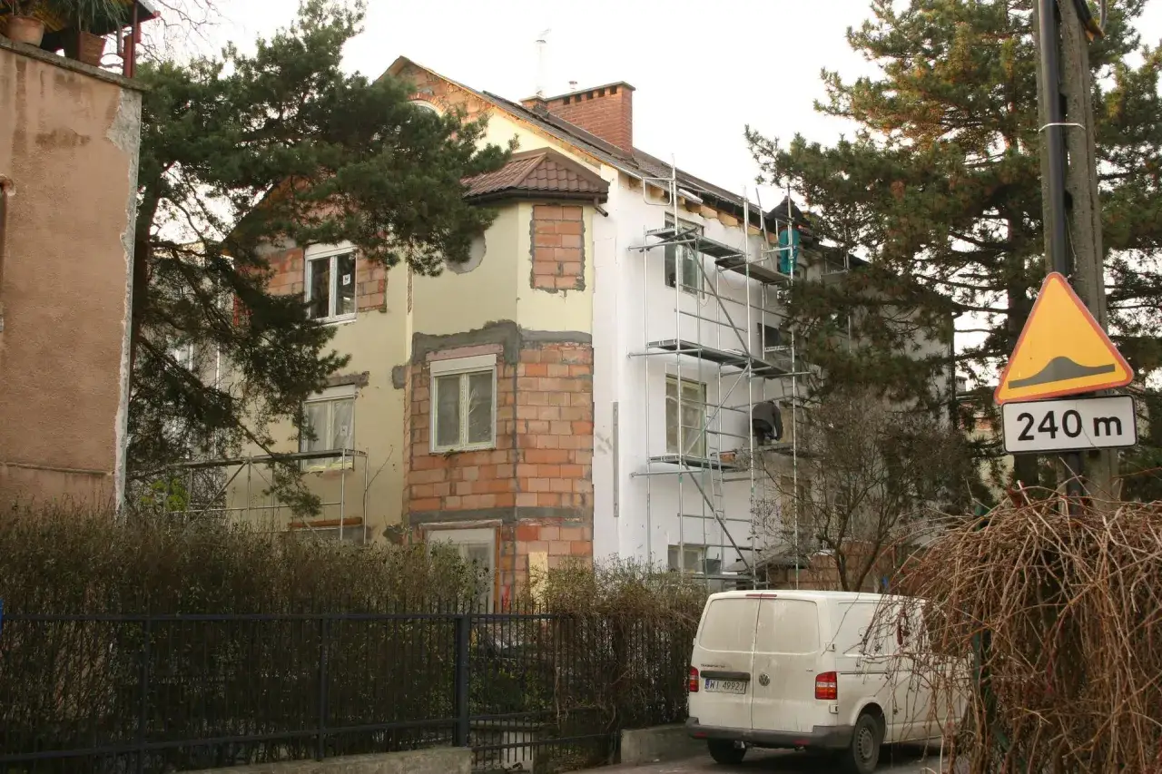 Trwa remont starego domu. Widać rusztowania, styropian i cegły. Obok stoi biały van.
